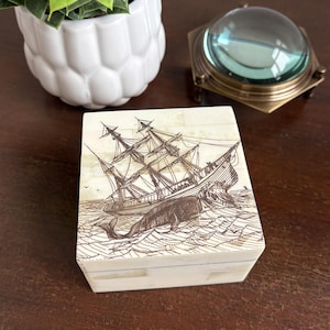 May include: A square, off-white box with a detailed illustration of a sailing ship and a whale in a sea. The artwork is in shades of brown. The box is likely for storing small items.