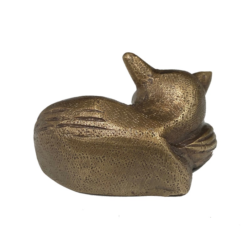 May include: A small, gold-colored metal sculpture of a cat curled up sleeping. The sculpture has a detailed, textured surface.