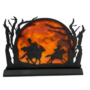May include: Black silhouette of two figures riding horses against a backdrop of a large orange moon and trees. The moon has a textured, marbled appearance.