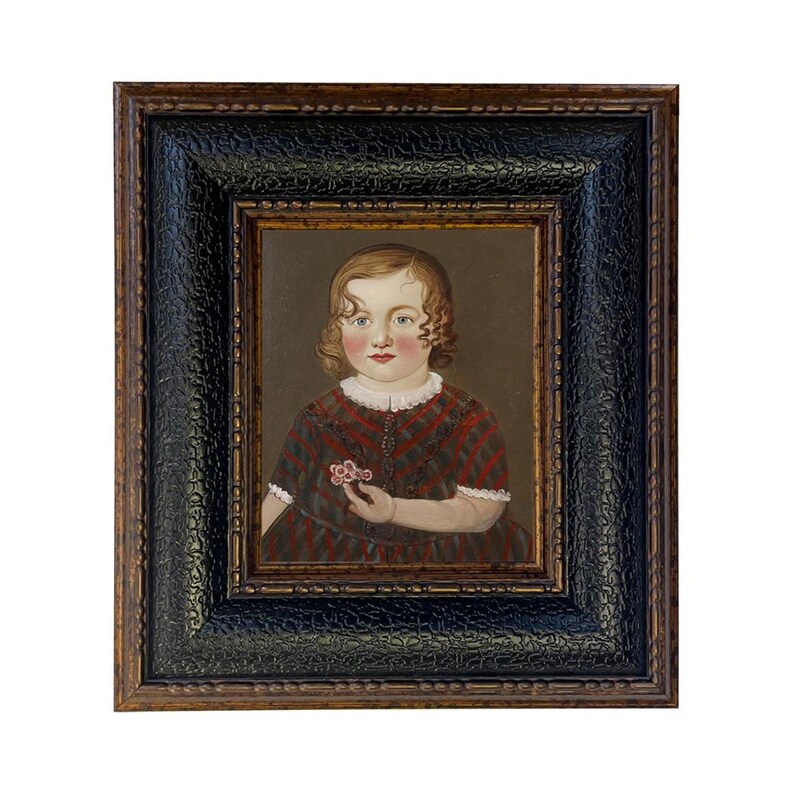 May include: An antique portrait of a young child in a dark wooden frame. The child is wearing a red and black plaid dress and holding a small flower. The frame has a textured black border and a gold inner edge.