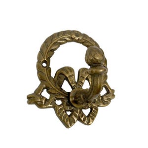 Könnte beinhalten: Ein dekorativer Wandhaken aus Messing mit einem verzierten Design. Der Haken hat einen kreisförmigen Kranz mit Blattdetails und einer stilisierten Schleife. Der Haken selbst ist gebogen und erstreckt sich von einer floralen Basis, ideal zum Aufhängen von Gegenständen.