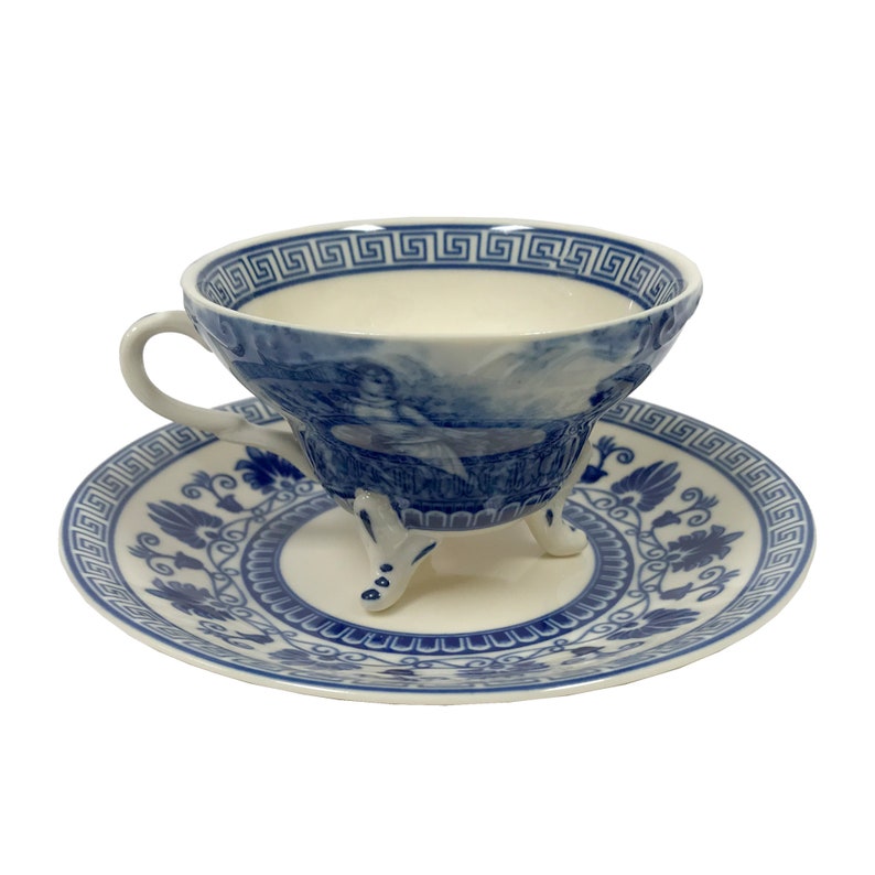 May include: A blue and white porcelain teacup and saucer set with a Greek key design. The teacup has three legs and features a blue and white scene of a woman in a landscape.