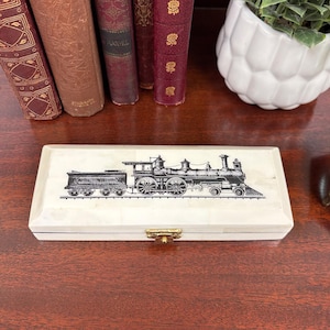 May include: A rectangular, off-white box with a black and white illustration of a steam train. The box has a gold-colored clasp and is placed on a dark wooden surface. The box is surrounded by books and a white pot with a plant.