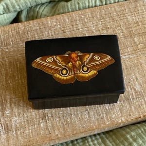 May include: A black rectangular box featuring a detailed moth illustration on the lid. The moth has brown and yellow wings with intricate patterns and eye-like markings. The box is on a light-coloured wooden surface.