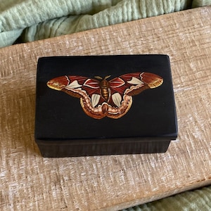 Puede incluir: Una caja de madera negra con una imagen pintada de una mariposa roja, blanca y marrón. La mariposa tiene un diseño detallado y está centrada en la tapa de la caja.