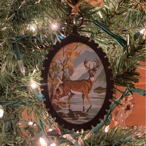 May include: A brown metal Christmas ornament with a painted image of a deer in a forest scene. The ornament is oval shaped and has a scalloped edge.