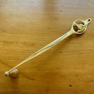 May include: A gold-toned candle snuffer with a long, thin handle and a decorative head featuring a dog's face. The snuffer is on a wooden surface.