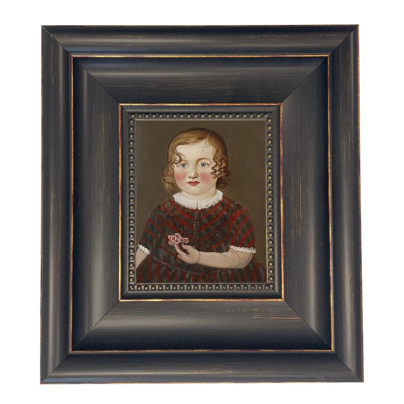 May include: A framed portrait of a young child with curly hair, wearing a red and brown plaid dress, holding a small flower. The portrait is set within a dark, ornate wooden frame with gold accents, creating a vintage aesthetic.
