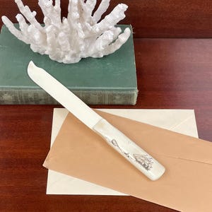 May include: A letter opener with a smooth, off-white blade and a matching handle featuring a detailed black ink illustration of a sailing ship. The handle is cylindrical and tapers slightly toward the blade, showcasing a nautical design.