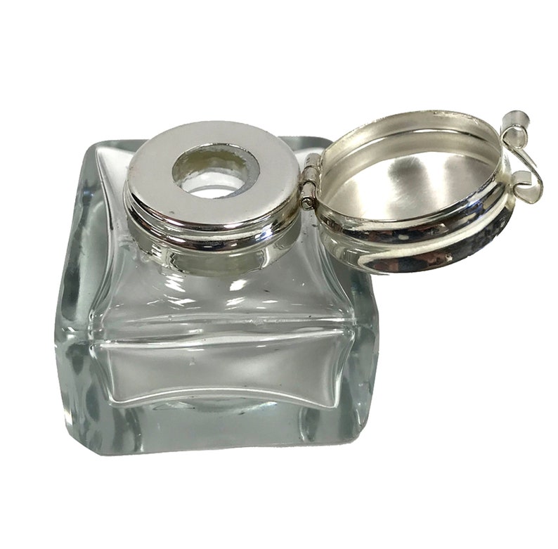 May include: A clear glass inkwell with a silver hinged lid. The lid has a round opening in the center.