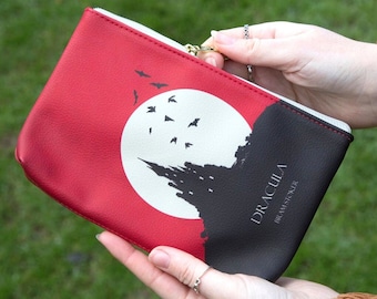 Pode incluir: Uma bolsa com zíper vermelha e preta com uma silhueta de lua e castelo brancos, morcegos voando ao redor da lua. O texto "DRACULA" e "BRAM STOKER" está impresso na bolsa.