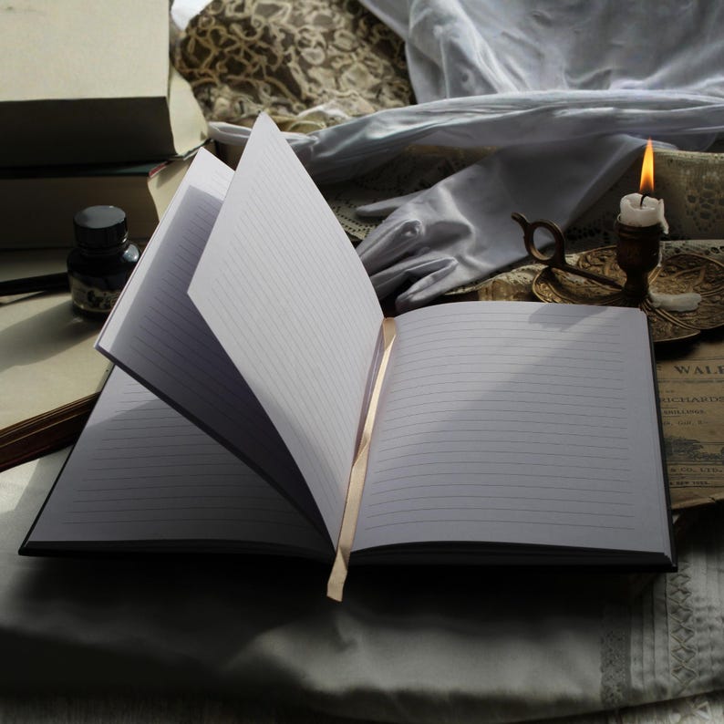 May include: An open, lined notebook with a gold ribbon bookmark. The pages are crisp white, and the cover is black. A lit candle, inkwell, and other vintage items are in the background, creating a classic, literary aesthetic.