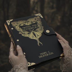 Puede incluir: Un diario negro con detalles dorados y el título "Moths and Butterflies" en letras doradas. El diario presenta un diseño de mariposa dorada y el nombre del autor, Mary C. Dickerson, también está en oro. El diario tiene un cierre a presión.