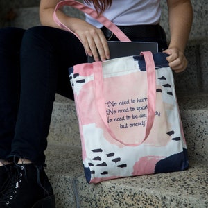 May include: A pink tote bag with a white background and abstract pink, blue, and black watercolor-style designs. The bag has a quote printed on it that reads: "No need to hum No need to spar No need to be any but oneself."