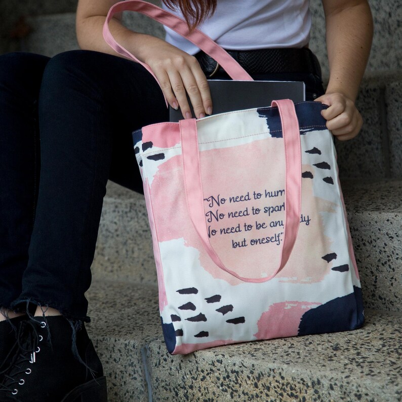 May include: A pink tote bag with a white background and abstract pink, blue, and black watercolor-style designs. The bag has a quote printed on it that reads: "No need to hum No need to spar No need to be any but oneself."