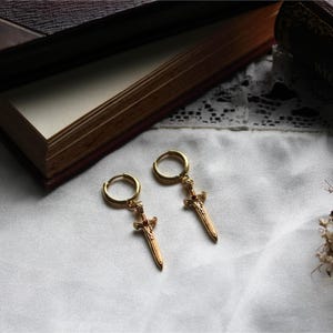 May include: A pair of gold-colored hoop earrings with sword pendants. Each earring features a small red gem at the top of the sword. The earrings are displayed on a white surface, with a book in the background.