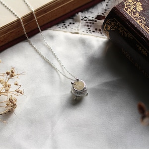 Puede incluir: Collar plateado con un peque&ntilde;o colgante de caldero ornamentado. El colgante tiene un interior claro y detalles decorativos en el borde y la base. La delicada cadena descansa sobre una tela blanca.