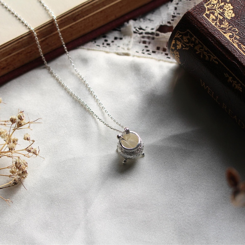 Puede incluir: Collar plateado con un peque&ntilde;o colgante de caldero ornamentado. El colgante tiene un interior claro y detalles decorativos en el borde y la base. La delicada cadena descansa sobre una tela blanca.