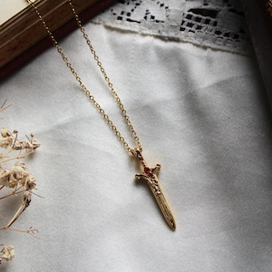 Puede incluir: Un collar con colgante dorado con un diseño de espada con una gema roja en la empuñadura. El colgante de espada cuelga de una delicada cadena. El collar se muestra sobre una superficie de tela blanca.
