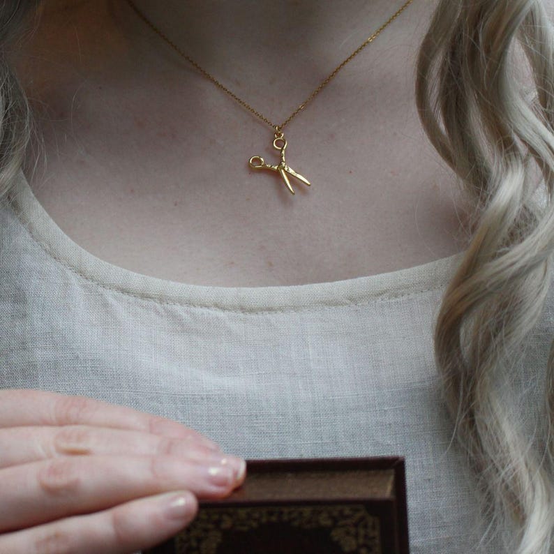 Peut inclure: Un collier en or avec un pendentif en forme de ciseaux. Le collier est port&eacute; par une personne aux longs cheveux blonds.