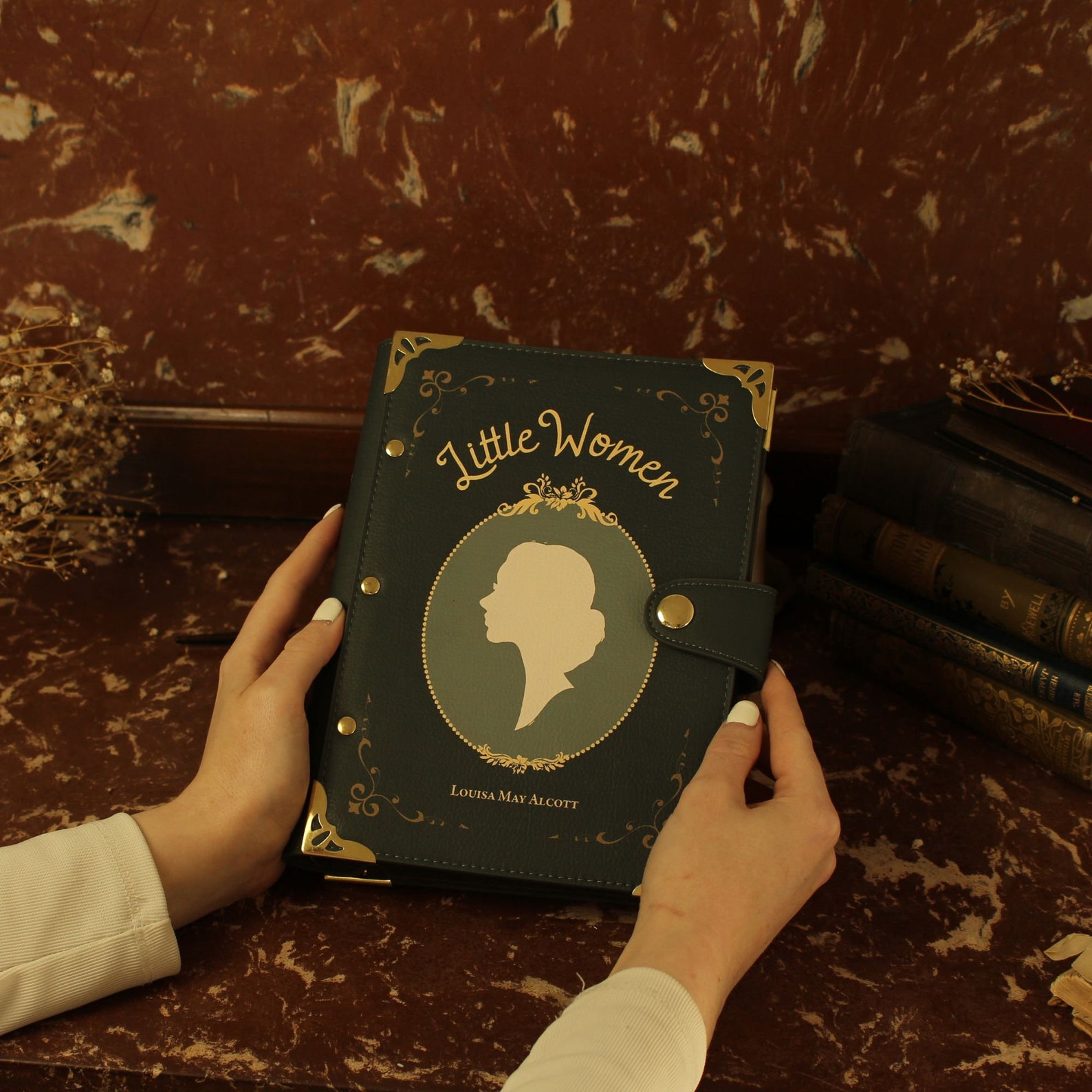 A marbled maroon and tan desk and wall with a white person holding a green notebook. It has gold corners and a clasp and says "Little Women" on it above a framed silhouette of a woman.