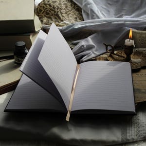 May include: An open, lined notebook with a gold ribbon bookmark. The pages are crisp white, and the cover is black. A lit candle, inkwell, and other vintage items are in the background, creating a classic, literary aesthetic.