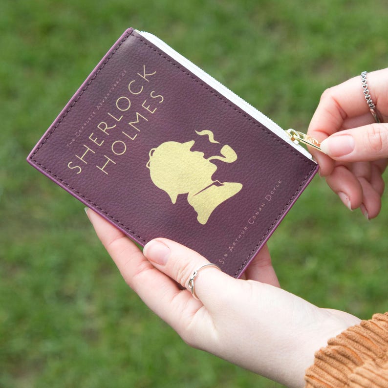 May include: A deep plum-colored zippered pouch with a white zipper and gold zipper pull. The pouch is styled like a book cover and features gold foil text reading 'The Complete Adventures of Sherlock Holmes' by Sir Arthur Conan Doyle. A gold silhouette of Sherlock Holmes with a deerstalker hat and pipe is prominently displayed on the front. The pouch is held in both hands against a grassy outdoor background.