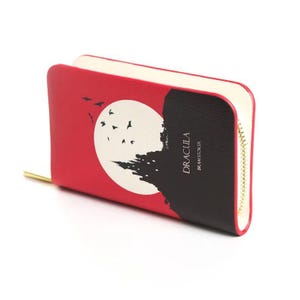May include: A red and black wallet featuring a design inspired by Bram Stoker's Dracula. The design includes a white full moon, a silhouette of a castle, and flying bats. The wallet has a gold zipper and the word "Dracula" is printed on the front.
