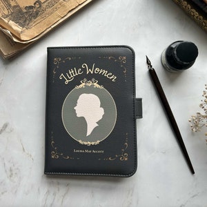 Può includere: Un libro rilegato in pelle nera con un titolo in rilievo dorato "Piccole Donne" e una silhouette di una testa di donna. Il nome dell'autore, Louisa May Alcott, è stampato in oro sotto la silhouette.