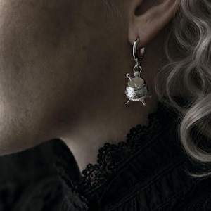 May include: A silver-colored earring featuring a miniature cauldron design. The cauldron has three small legs and a lid, with a spoon inside. The earring is attached to a silver hoop. The image is a close-up, showing the earring in detail.