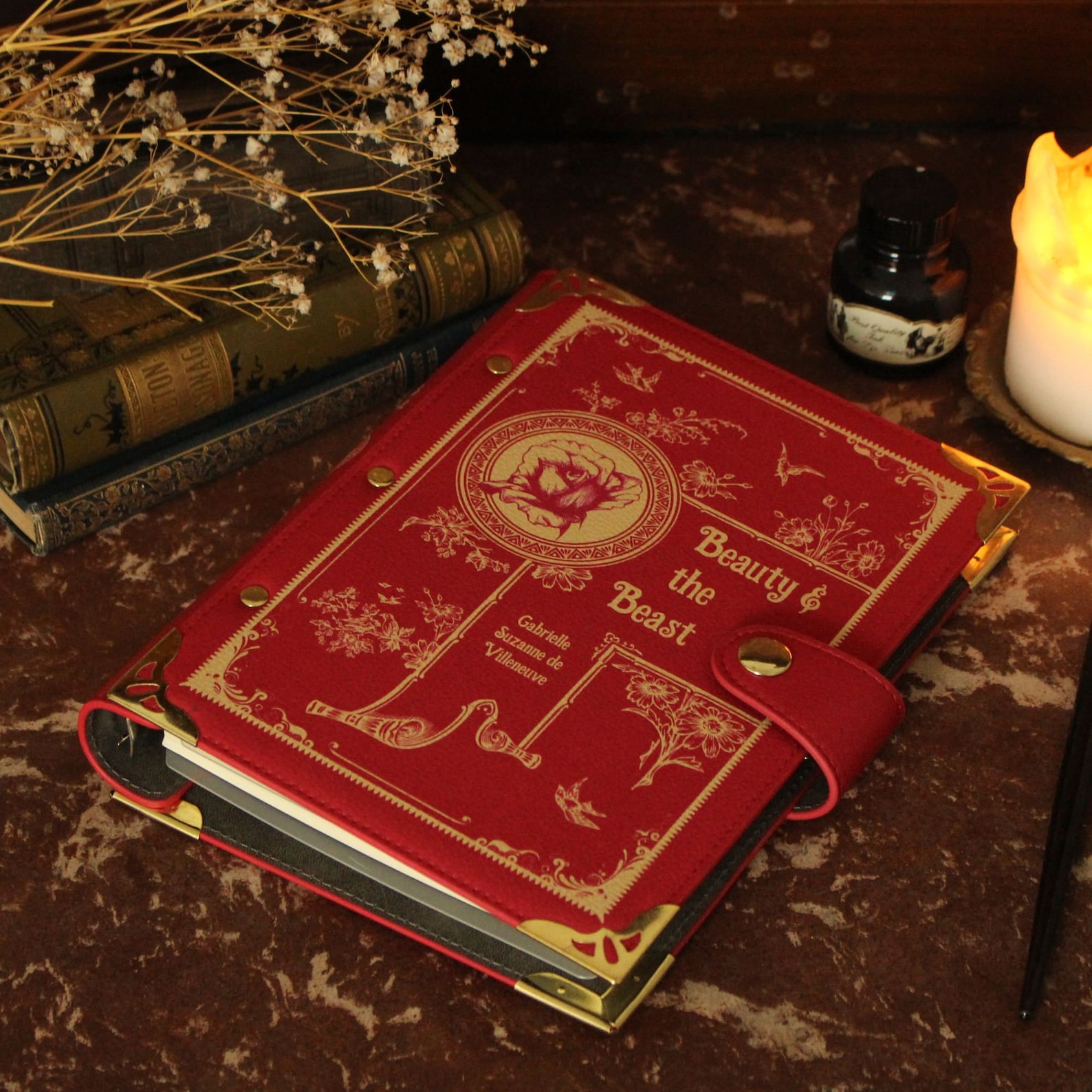A marbled tan and brown desk with a red leather binder on it. It says "Beauty & the Beast" on the front with a wax seal-like design with a rose in it, and other ornate gold designs.