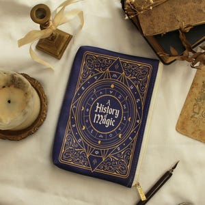 May include: A navy blue zippered pouch in the shape of a book, featuring gold-coloured decorative patterns. The text "A History of Magic" is displayed on the front. A gold zip and a pen are also visible.