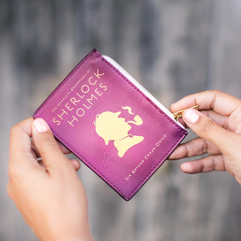 Puede incluir: Una bolsa de cremallera morada con un dise&ntilde;o de portada de libro que presenta el texto "The Complete Adventures of Sherlock Holmes" y una ilustraci&oacute;n de Sherlock Holmes. El texto "Sir Arthur Conan Doyle" est&aacute; impreso en la parte inferior de la bolsa.