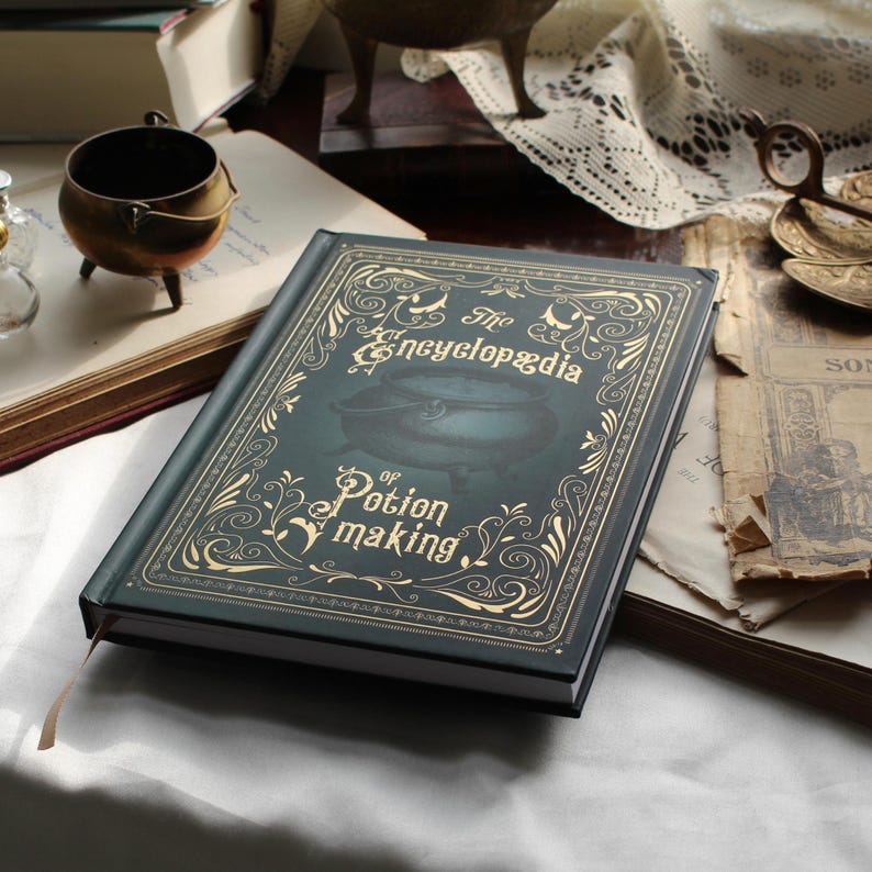 May include: A dark green book titled "The Encyclop&aelig;dia of Potion Making" with gold lettering and illustrations. The book has a decorative cover with a cauldron image. A bronze cauldron and open books are in the background.
