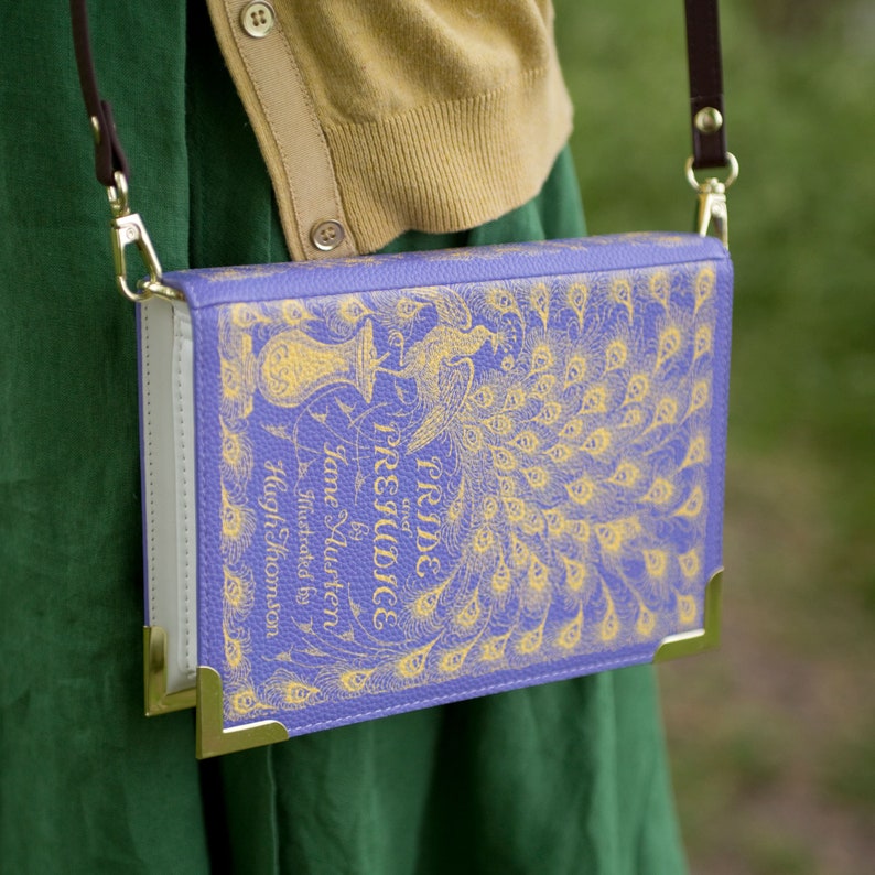 May include: A purple book bag with a gold clasp and a brown strap. The bag features a peacock design and the text "Pride and Prejudice by Jane Austen Illustrated by Hugh Thomson".