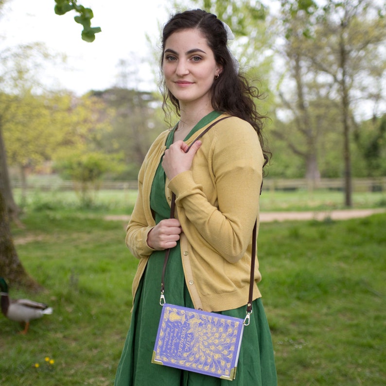 May include: A woman wearing a green dress and a yellow cardigan is holding a book-shaped purse with a peacock design. The purse has a brown strap and is worn over the shoulder.