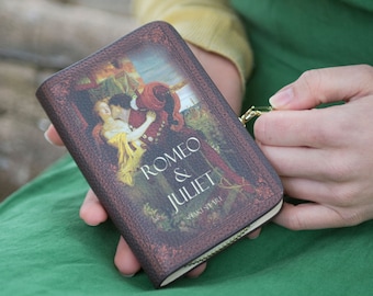 Könnte beinhalten: Eine kleine, rechteckige Geldbörse mit dem Cover von Shakespeares "Romeo & Julia". Das Design beinhaltet eine romantische Illustration der Charaktere und den Titeltext. Die Geldbörse hat ein braunes, strukturiertes Aussehen und einen goldenen Reißverschluss.