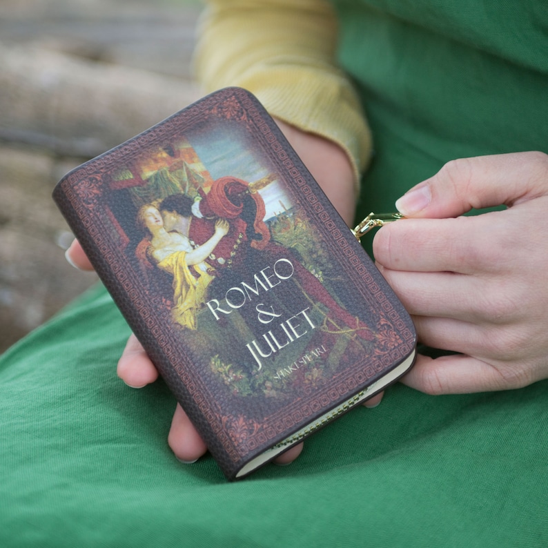 May include: A small, rectangular wallet featuring the cover art of Shakespeare's "Romeo & Juliet." The design includes a romantic illustration of the characters and the title text. The wallet has a brown, textured appearance and a gold zip.