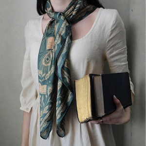 Può includere: Una sciarpa di seta verde acqua con un motivo di piume di pavone e libri è drappeggiata intorno al collo. L'outfit è completato da un abito color crema. Due libri antichi con bordi dorati sono tenuti in mano.