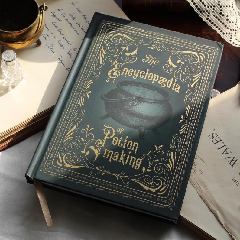 May include: A dark green book titled "The Encyclopedia of Potion Making" with gold lettering and a cauldron illustration. The book is open, revealing a cream-colored bookmark. The cover has ornate gold filigree.