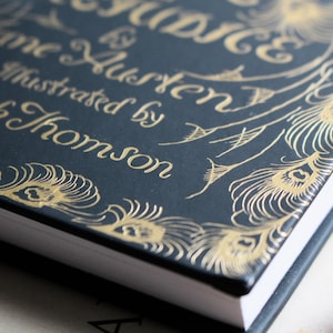May include: Close-up of a book cover with the title "Pride & Prejudice" by Jane Austen, illustrated by Hugh Thomson. The cover is dark blue with gold lettering and peacock feather designs.