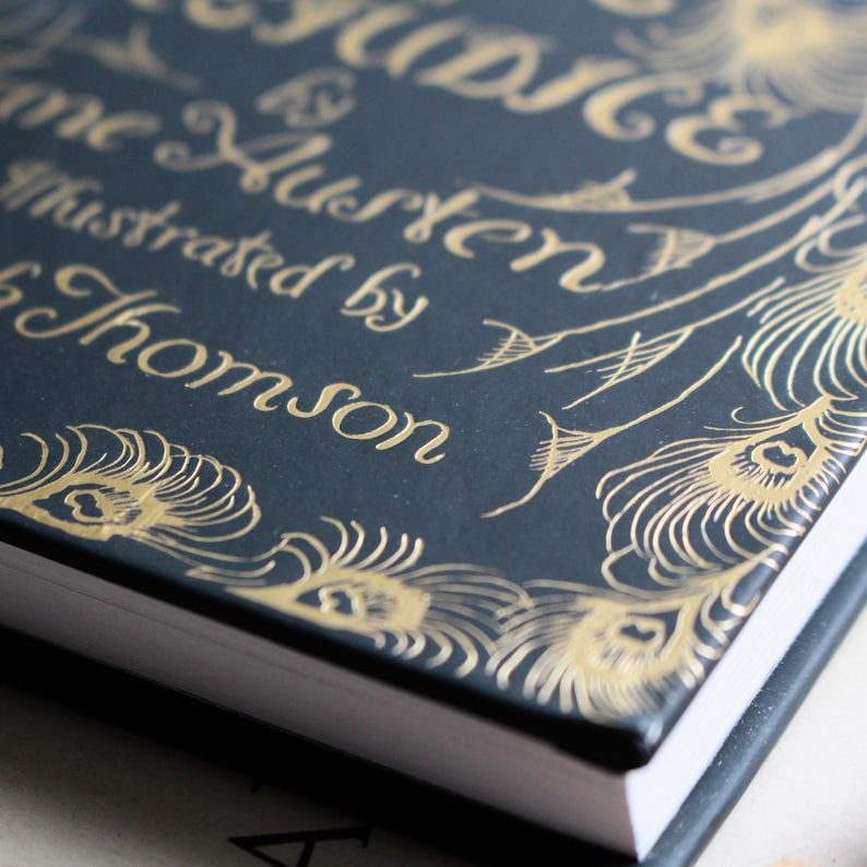 May include: Close-up of a book cover with the title "Pride & Prejudice" by Jane Austen, illustrated by Hugh Thomson. The cover is dark blue with gold lettering and peacock feather designs.