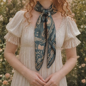 May include: A dark teal square scarf featuring a repeating pattern of open books, peacock feathers, and botanical elements in a light gold tone. The scarf is tied around the neck, worn over a cream-colored dress with short puff sleeves and lace trim.