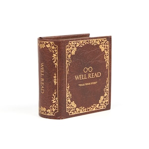 Peut inclure: Une bo&icirc;te en cuir marron en forme de livre avec des accents dor&eacute;s et le texte "WELL READ" et "WEAR YOUR STORY".