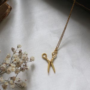 Peut inclure: Un collier en or avec un petit pendentif en forme de ciseaux d&eacute;taill&eacute;. Le collier est sur un fond blanc.