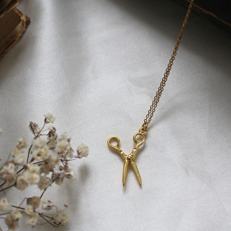Peut inclure: Un collier en or avec un petit pendentif en forme de ciseaux d&eacute;taill&eacute;. Le collier est sur un fond blanc.