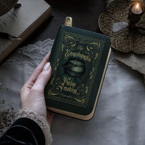 May include: A green, book-shaped case with gold detailing and the words "The Encyclopedia of Potion Making" on the cover. The case has a zip closure and a gold bookmark. The image has a vintage aesthetic, with a candle and open book in the background.