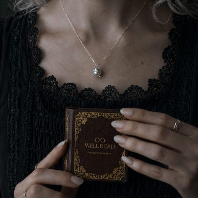 Puede incluir: Un collar de plata con un peque&ntilde;o colgante adornado cuelga sobre un top de encaje negro. Se sostiene un libro marr&oacute;n en miniatura con letras doradas que dice "WELL READ". La portada del libro tiene un borde dorado decorativo.