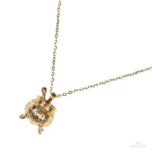 Puede incluir: Un collar dorado con un colgante detallado de caldero con dos peque&ntilde;as cucharas en su interior. El colgante tiene detalles ornamentados y est&aacute; suspendido de una delicada cadena. El collar se muestra sobre un fondo blanco.