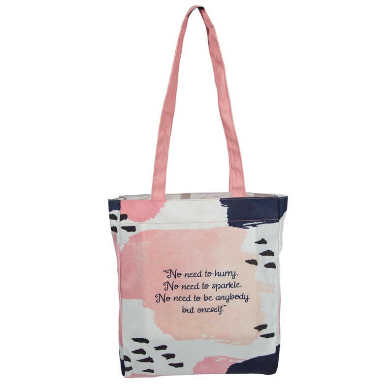 May include: A pink and white tote bag with black and blue abstract brushstrokes. The bag has pink straps and a quote printed on the front: "No need to hurry. No need to sparkle. No need to be anybody but oneself."